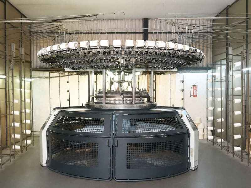 A large circular industrial knitting machine with numerous spools of white yarn