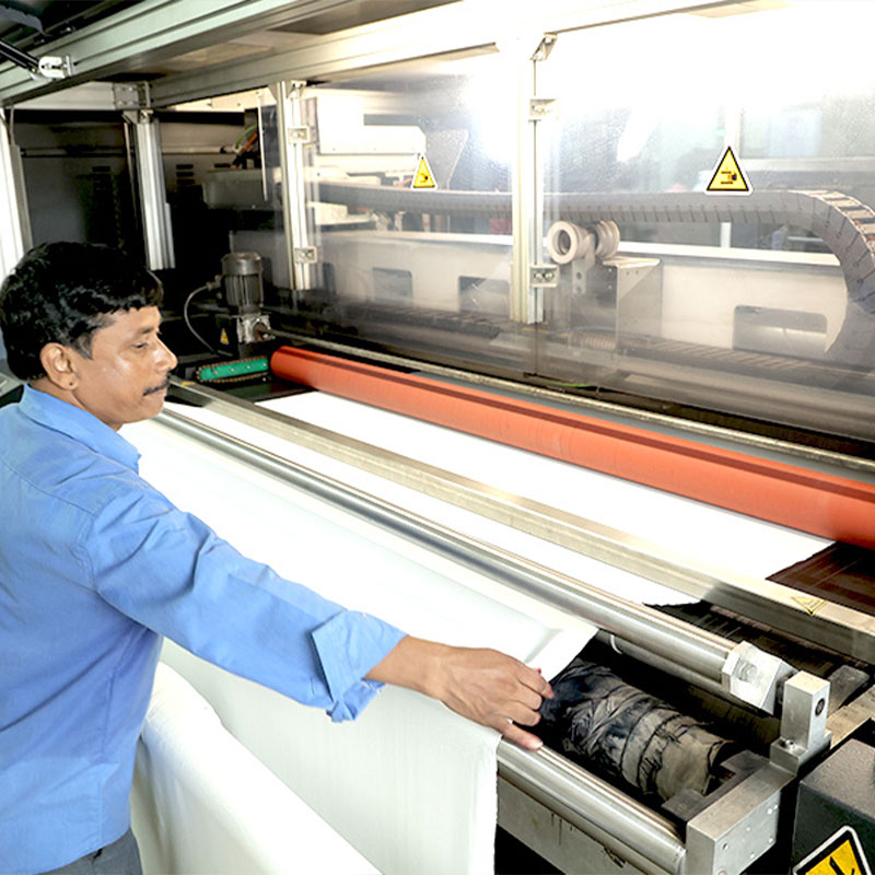 A worker in a blue shirt inspecting white fabric as it feeds through a large, industrial textile processing machine with an orange roller.